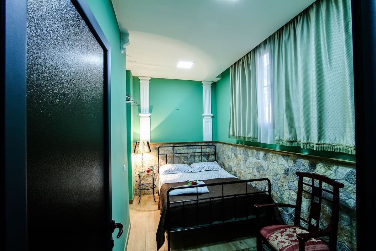 Une chambre à coucher avec un lit, une chaise, une table de chevet et une fenêtre avec des rideaux.