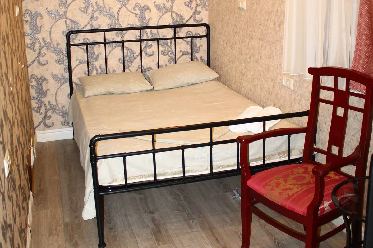 Un lit simple avec un cadre en métal, une chaise en bois avec un coussin rouge et un papier peint à motifs.