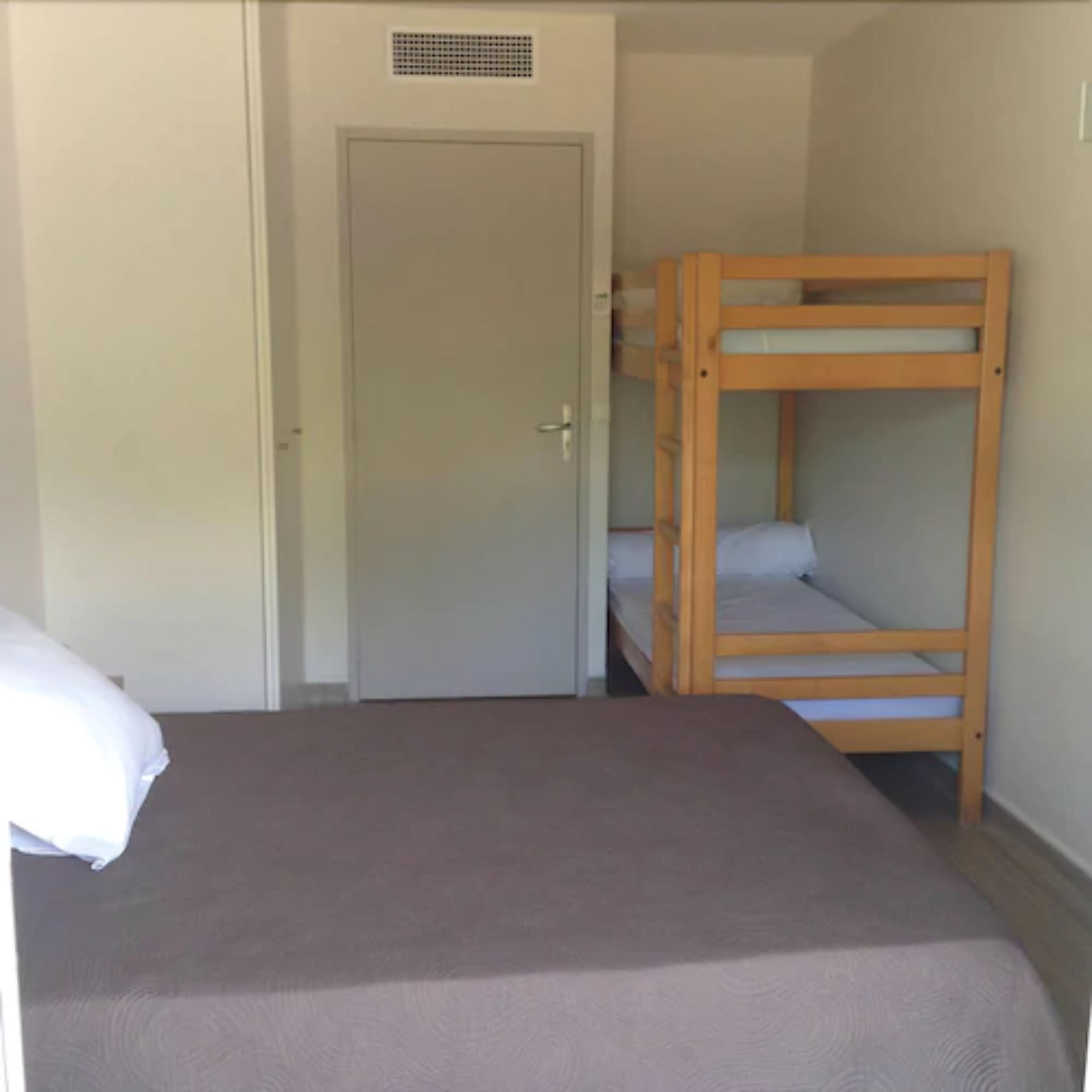 Een kamer met een eenpersoonsbed, een houten stapelbed en een deur met een ventilatieopening.