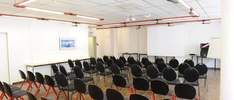 Un salón de conferencias con filas de sillas negras de patas rojas, paredes blancas y una pantalla de proyector.