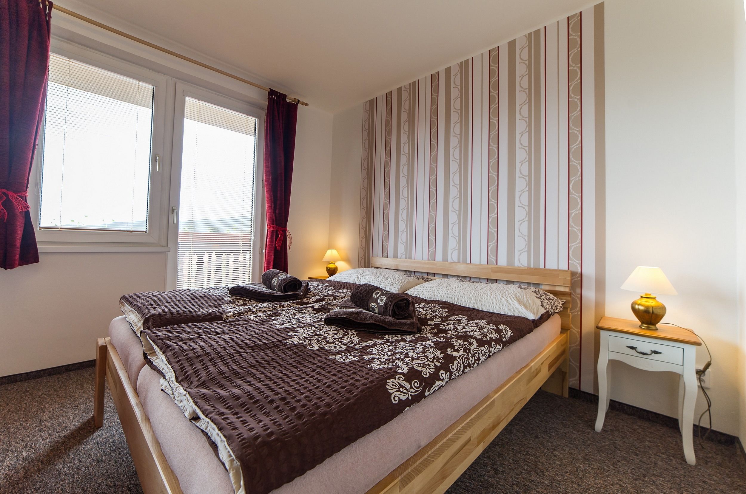 A neatly made bed with a patterned bedspread and two rolled towels, a bedside table with a lamp, and a window with curtains.
