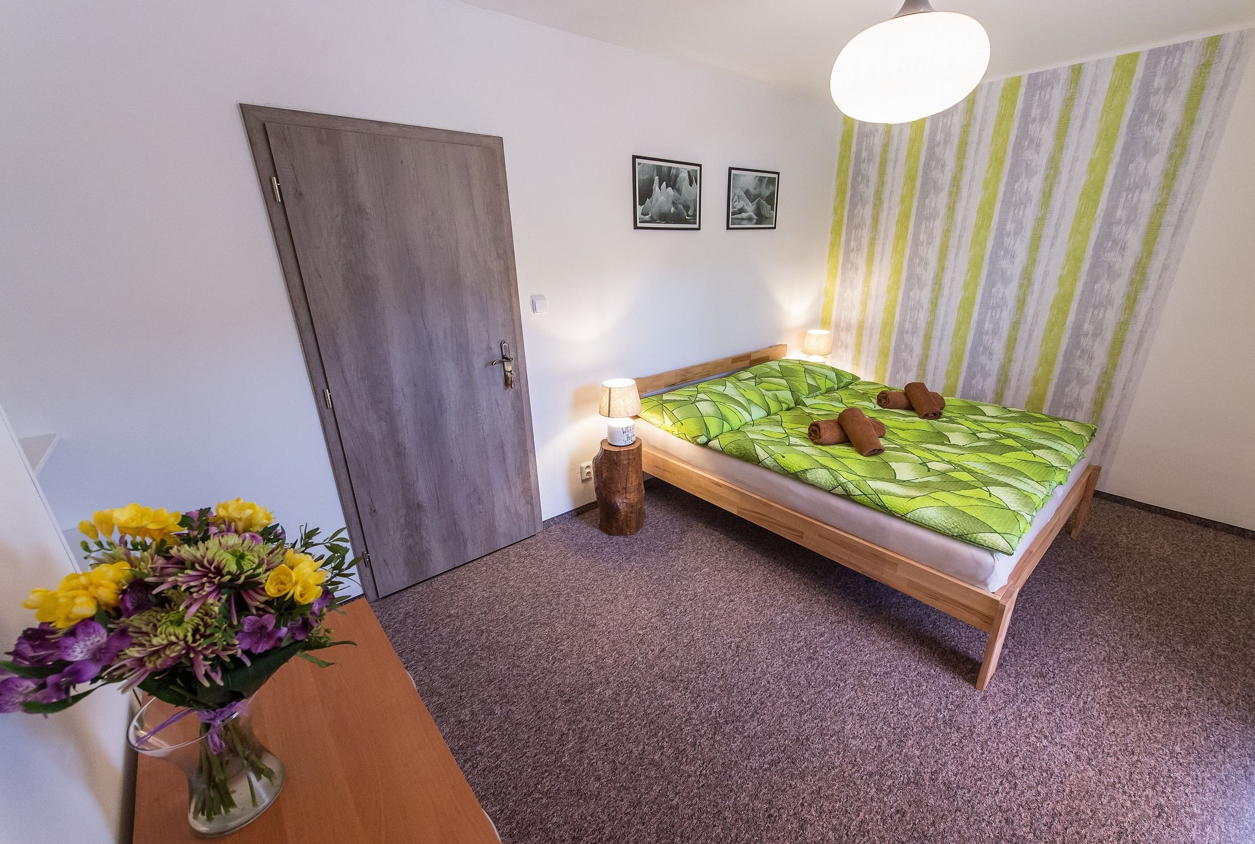 Un dormitorio con cama, una puerta de madera, un taburete pequeño de madera y un jarrón con flores.