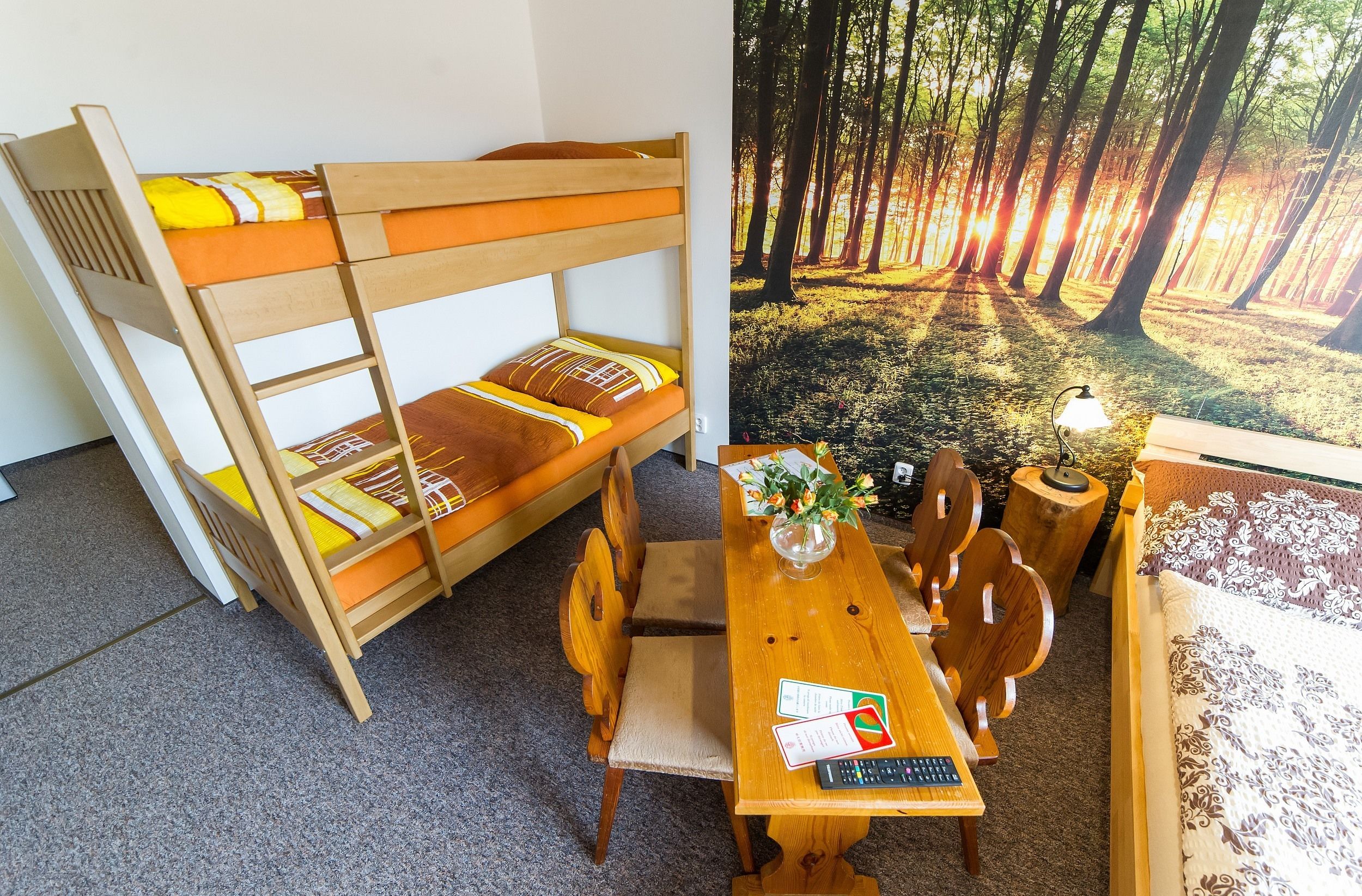 Uma cama de beliche com estrutura de madeira e roupa de cama laranja, uma mesa de jantar de madeira com cadeiras e uma pequena mesa de apoio com um abajur e flores.
