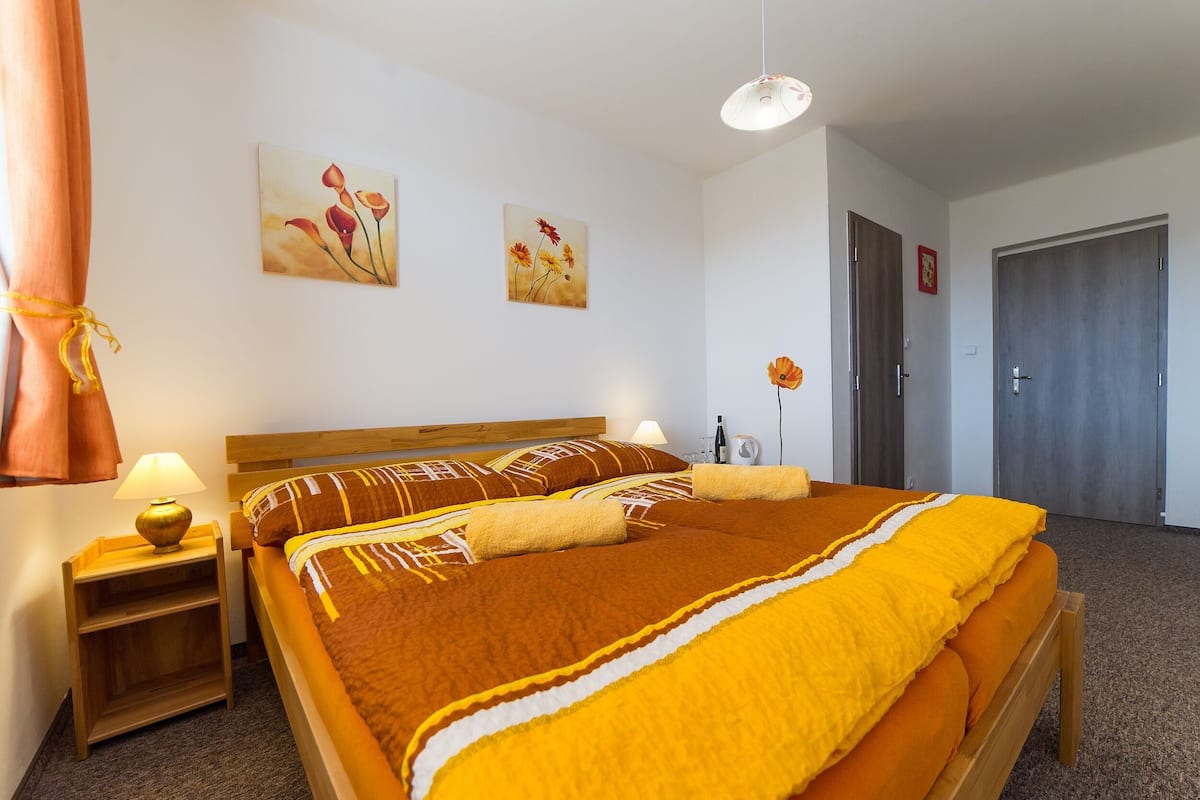 A hotel room with a large bed, wooden headboard, two wall-mounted paintings, a bedside table with a lamp, and a door to the right.