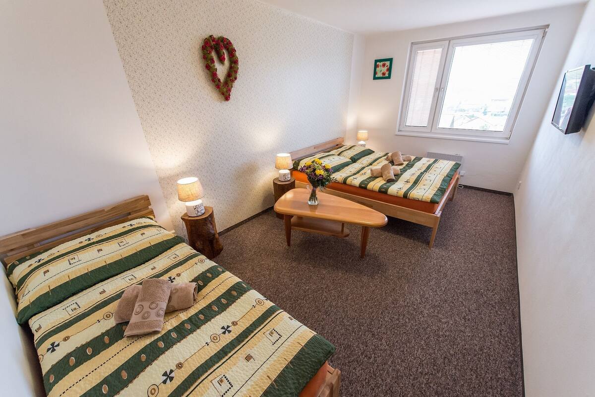 A hotel room with two beds, a wooden bench, a small table with a vase of flowers, and a window with blinds.