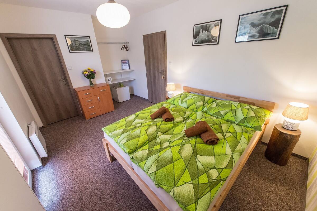 A bedroom with a bed, two towels, a wooden dresser, a small table with a lamp, and framed pictures on the wall.