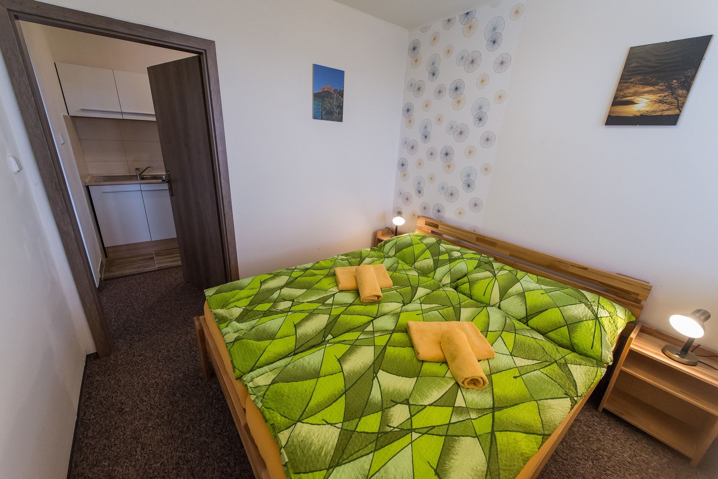A bedroom with a green patterned bedspread, two rolled towels, a nightstand with a lamp, and a view of a kitchen and bathroom through an open door.