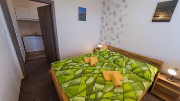 A bedroom with a green patterned bedspread, two rolled towels, a nightstand with a lamp, and a view of a kitchen and bathroom through an open door.
