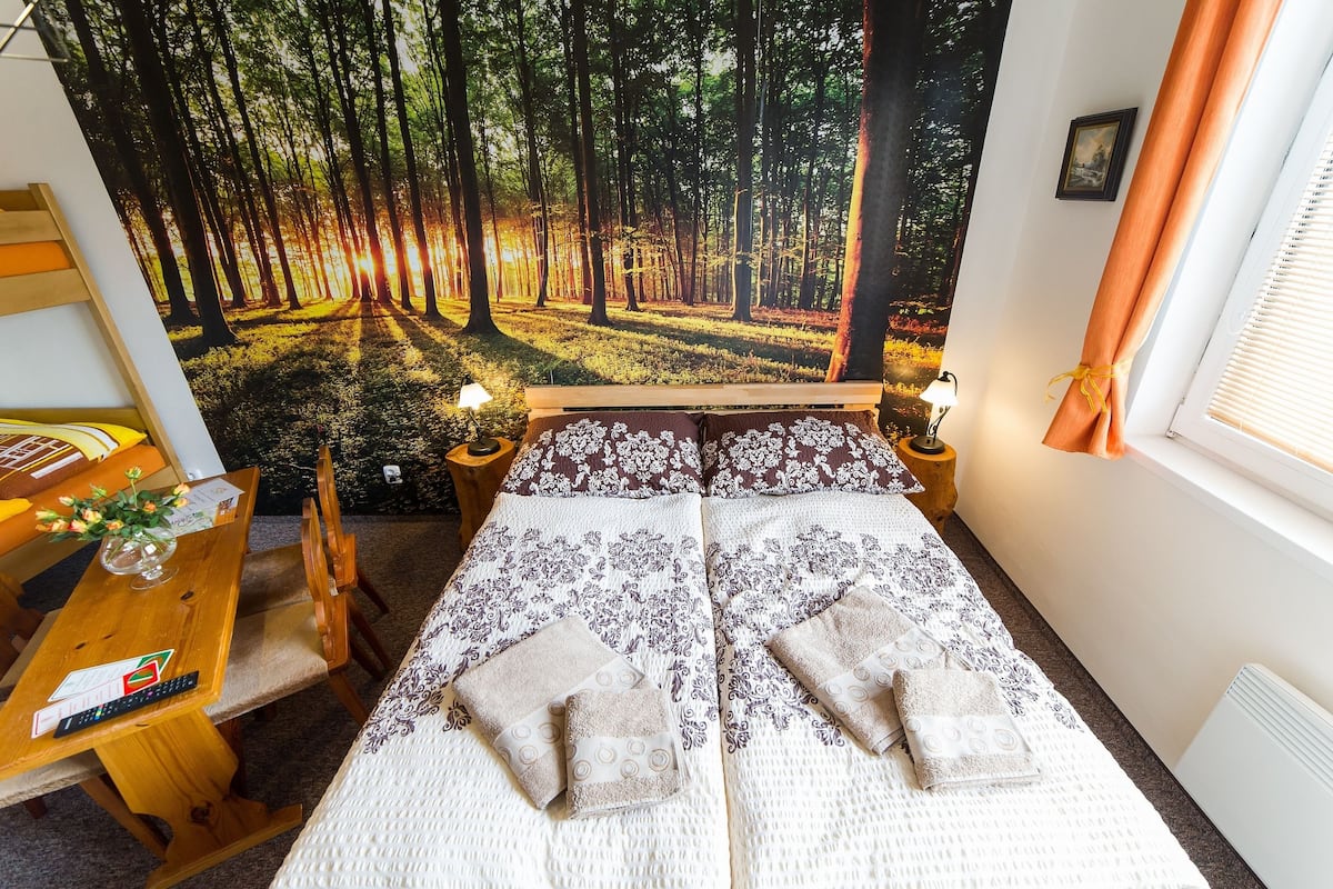 A bedroom with a large forest mural on the wall, a bed with patterned bedding, a wooden desk with a lamp, and a window with curtains.