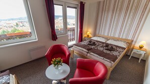 A hotel room with a bed, two red chairs, a small table with a vase of flowers, a window with a view of buildings, and a nightstand with a lamp. - Penzion Top (Cesky Krumlov)