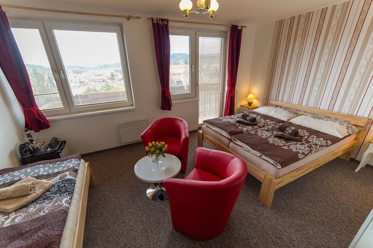 A bedroom with a bed, two red chairs, a small table with a vase of flowers, and a view of the city through large windows.