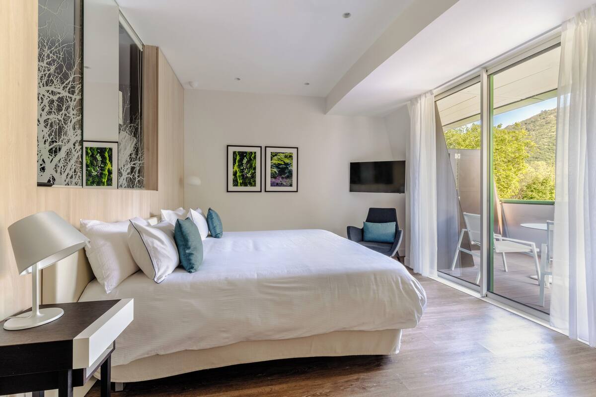 A modern bedroom with a large bed, a desk with a lamp, a TV, and a sliding glass door leading to an outdoor area.
