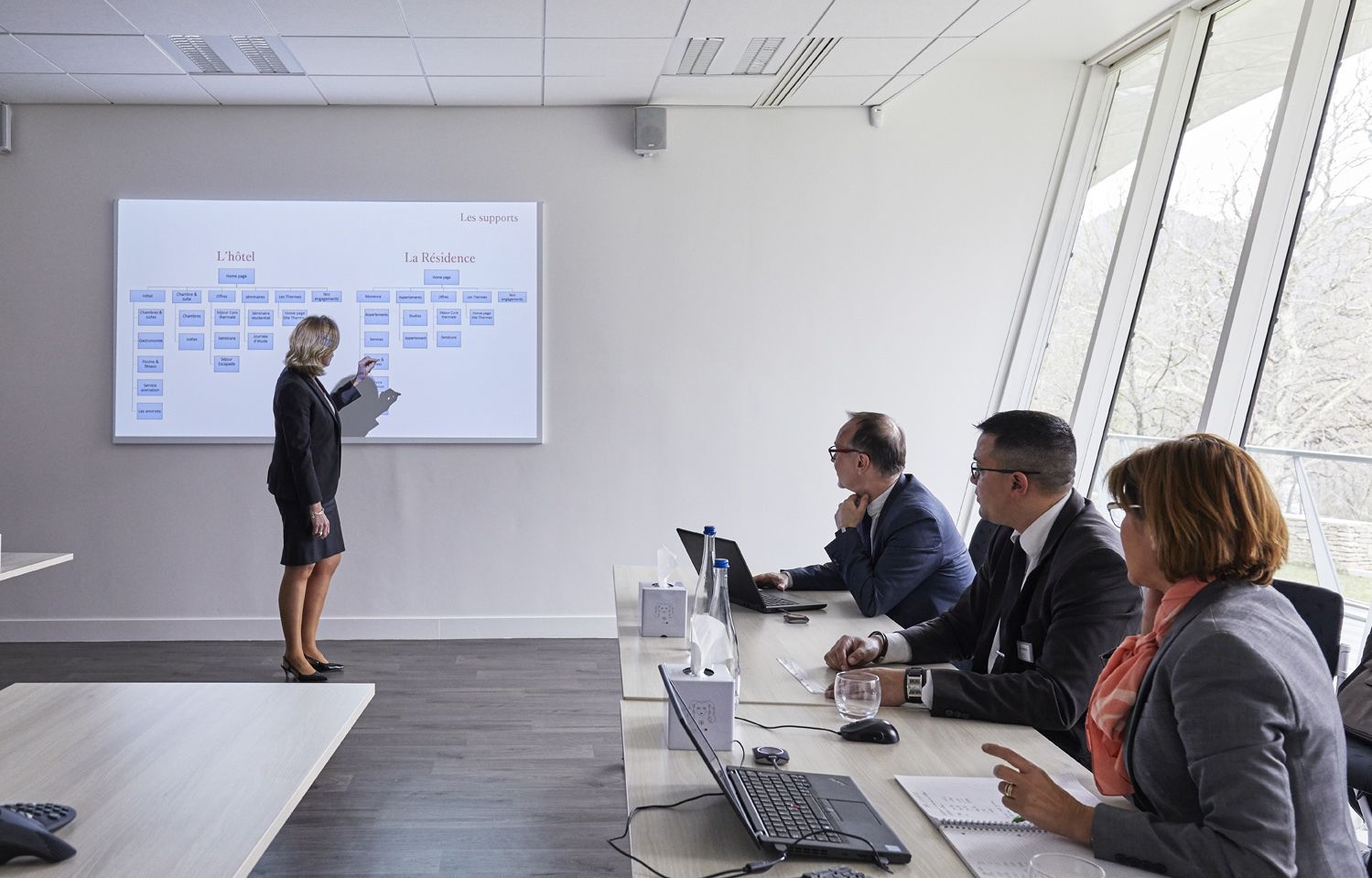 A business meeting in a modern conference room with a presentation on a screen, a long table with laptops, and a view of trees through large windows.