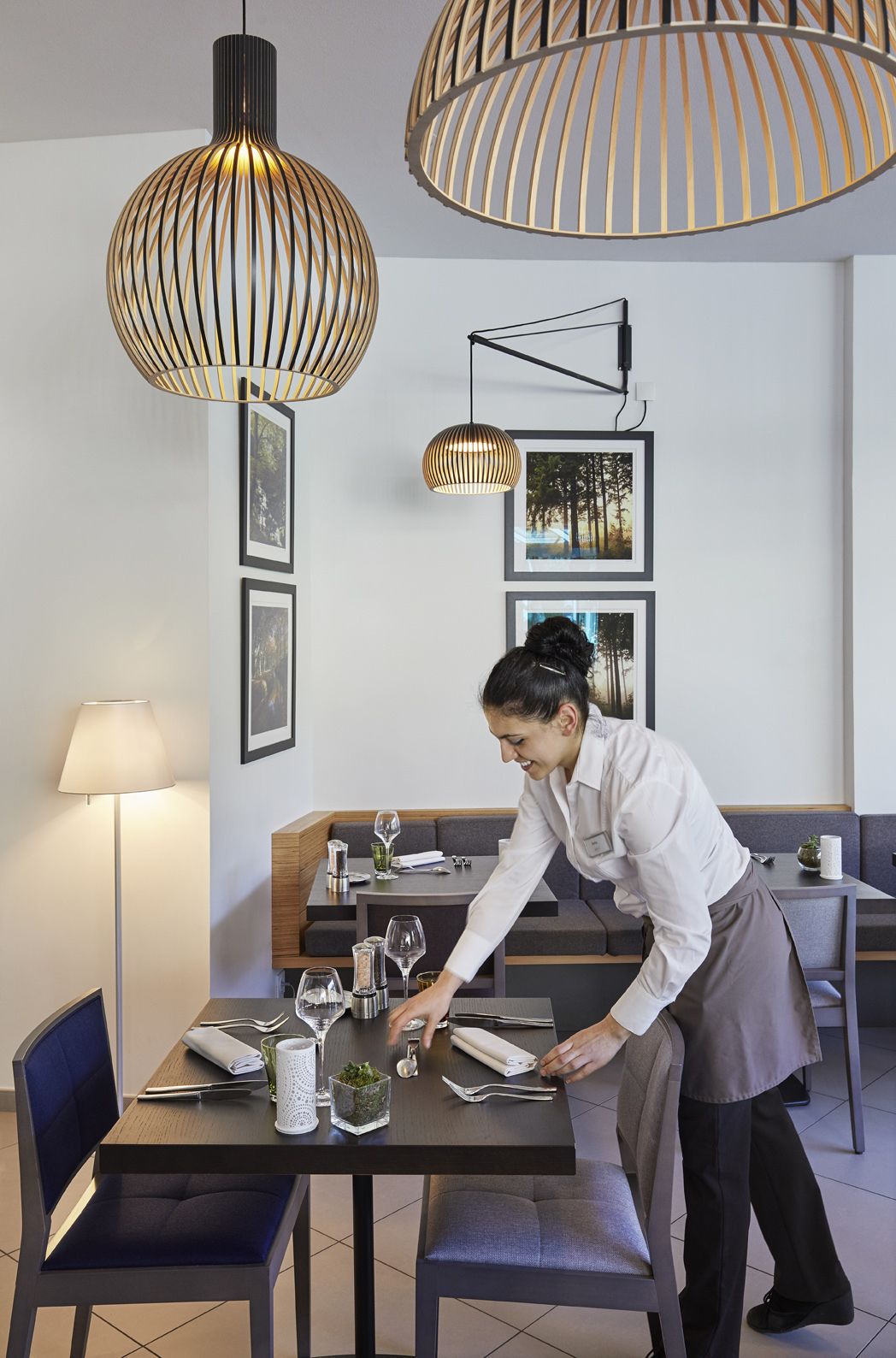 A modern dining area with a clean, minimalist design, featuring a table set for two with glasses and cutlery, and a person attending to the table.