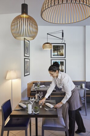 Un área de comedor moderna con un diseño limpio y minimalista, mostrando una mesa preparada para dos personas con vasos y cubiertos, y alguien atendiendo la mesa.