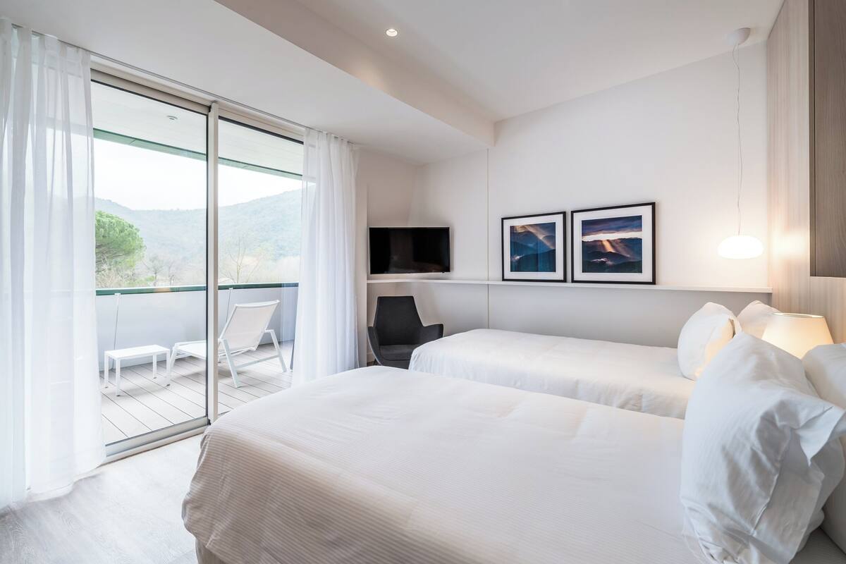A modern hotel room with two beds, a TV, a chair, and a balcony with a view of the mountains.