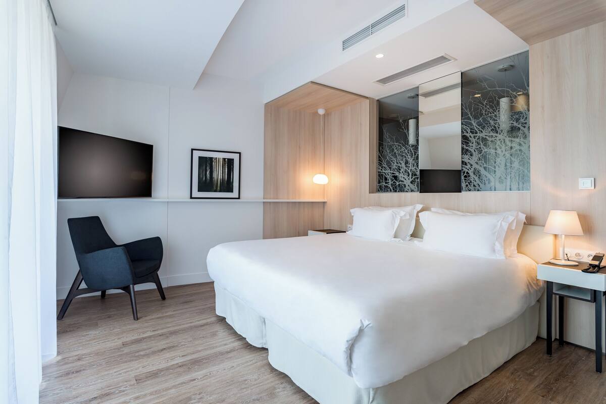 A modern hotel room with a large bed, a desk with a lamp, a chair, a flat-screen TV mounted on the wall, and a large mirror reflecting the room.