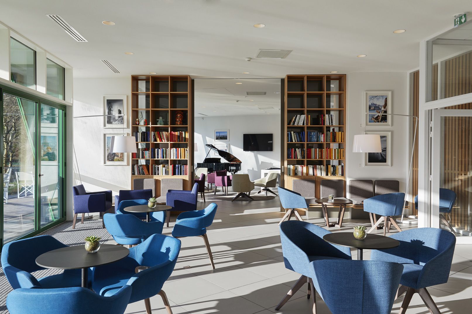 A modern hotel lobby with blue seating, large bookshelves, a grand piano, and a large window.