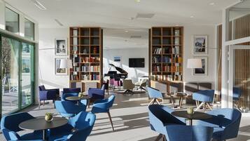 A modern hotel lobby with blue seating, large bookshelves, a grand piano, and a large window.