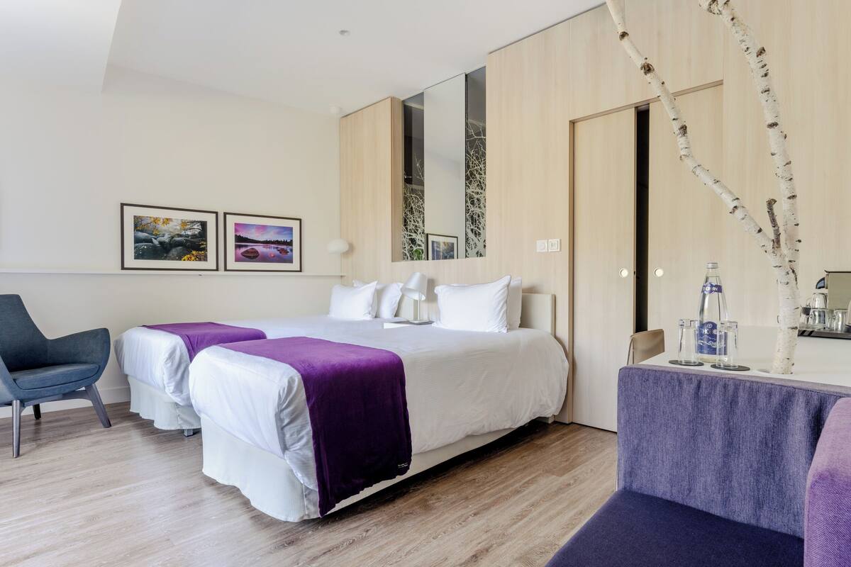 A modern hotel room with a large bed, two bedside tables, a chair, and a small table with a vase of branches.