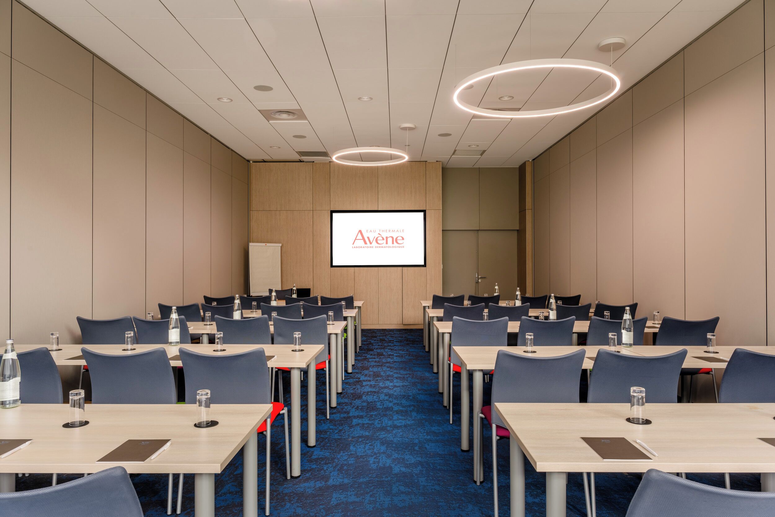 A conference room with a large screen displaying 'Eau de Vienne' branding, multiple tables with chairs, and a blue carpeted floor.