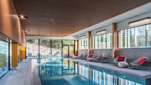 Piscina interior moderna com paredes de vidro transparente, teto de madeira e cadeiras confortáveis com almofadas vermelhas ao longo da borda.