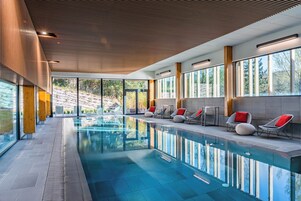 Una piscina interior moderna con paredes de vidrio transparente, techo de madera y sillones con cojines rojos a lo largo del borde.
