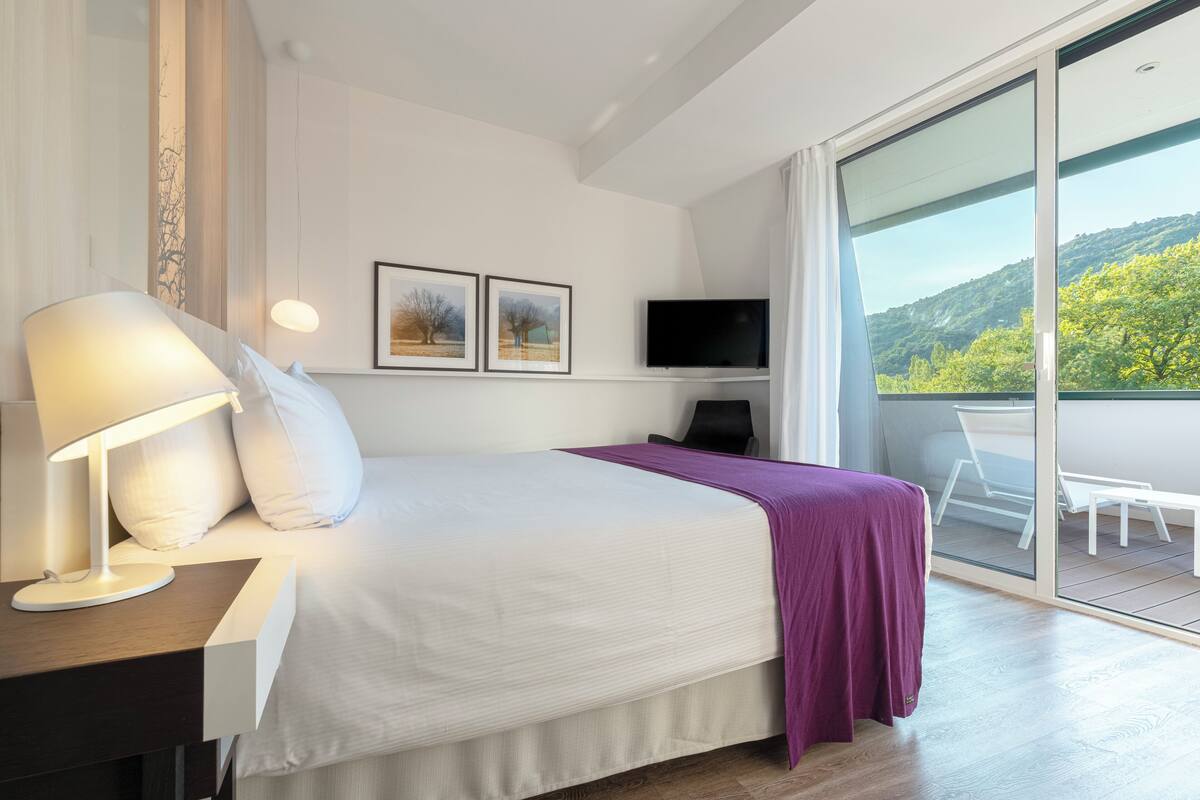 A modern hotel room with a bed, a desk with a lamp, a TV, and a balcony with a view of greenery.