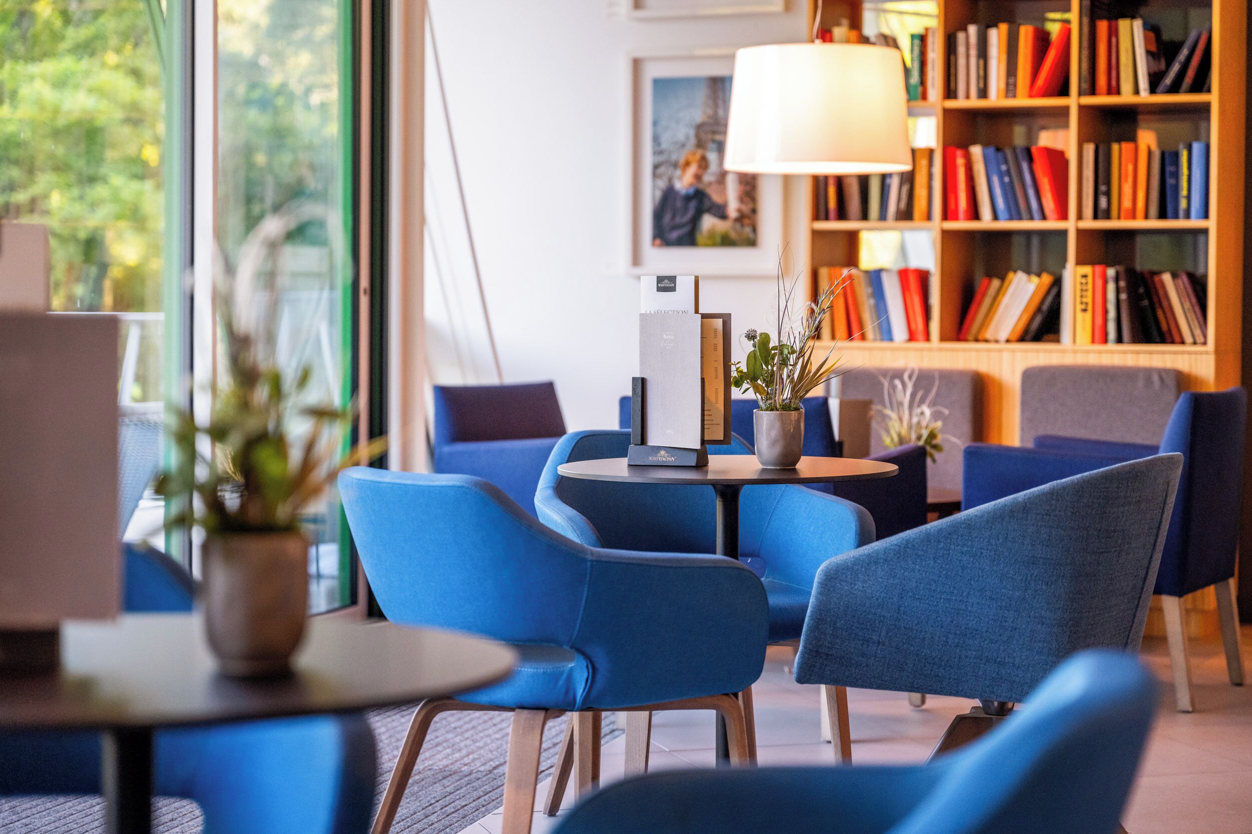 A modern lounge area with blue chairs, a small table, and a bookshelf filled with books.