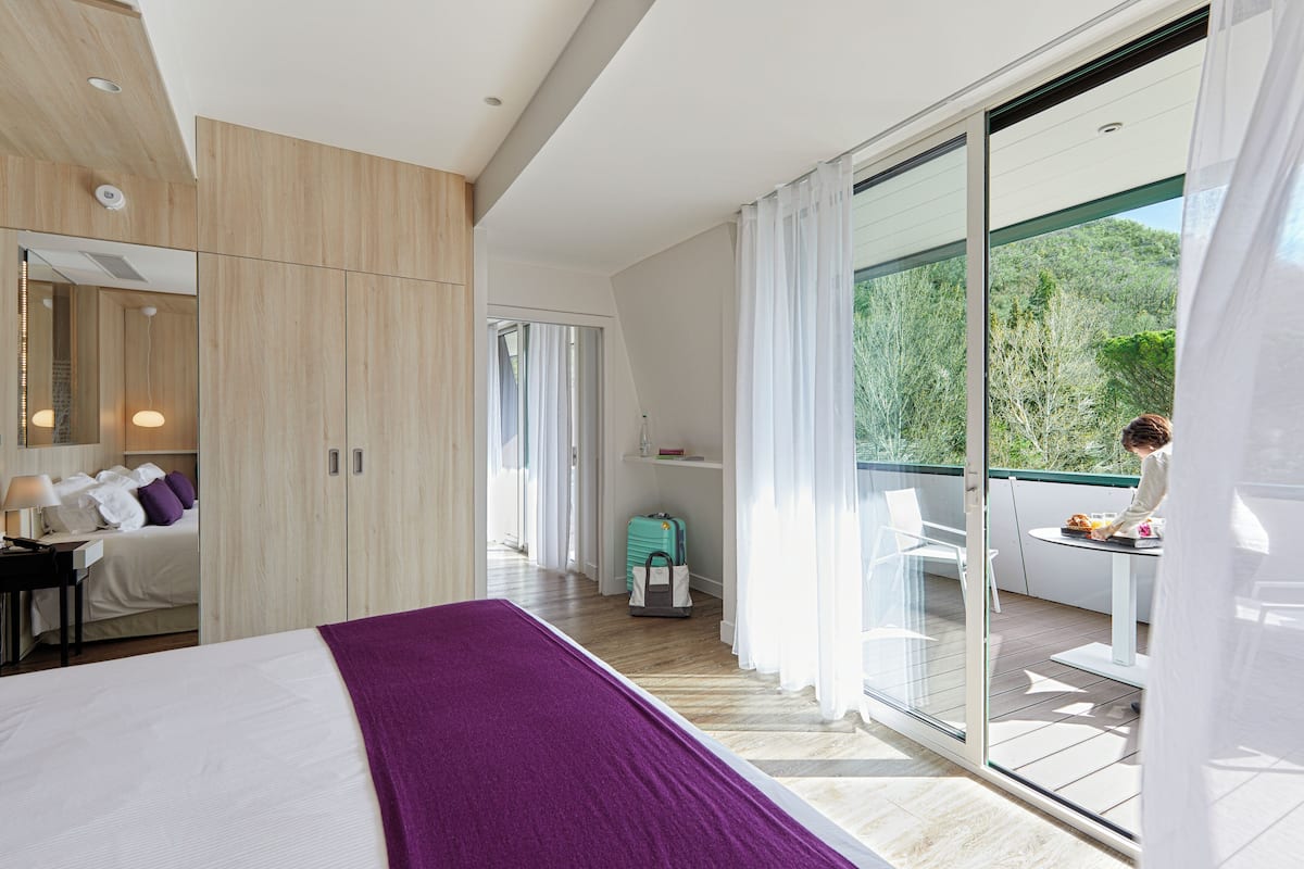 A modern hotel room with a large bed, a balcony with a table and chairs, and a view of greenery outside.