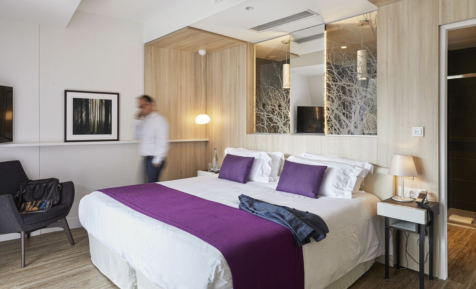 A modern hotel room with a large bed, a bedside table with a lamp, a chair, and a wall-mounted mirror.