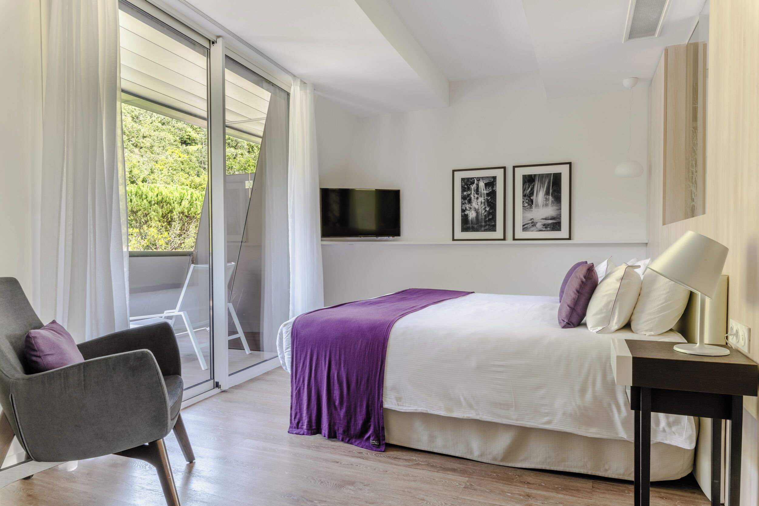A modern bedroom with a bed, a chair, a TV, and a window with curtains.