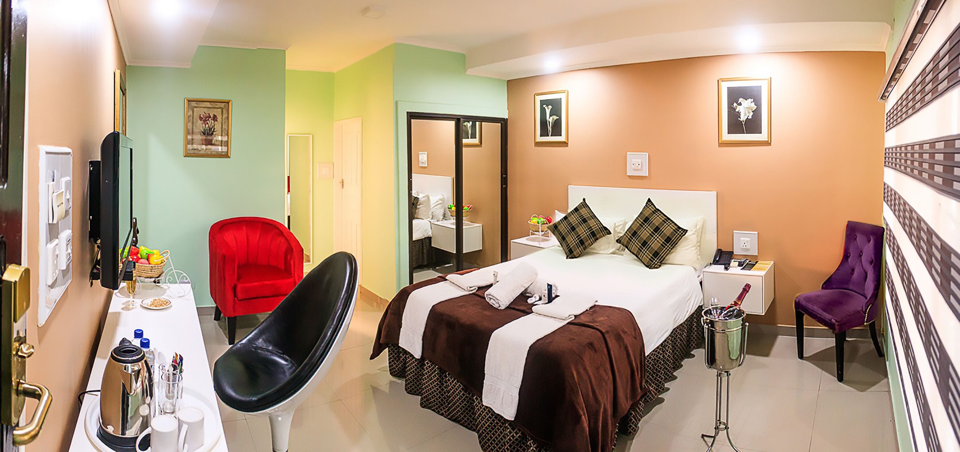 A hotel room with a bed, a red chair, a purple chair, a bedside table, a mirror, and a wall with framed pictures.