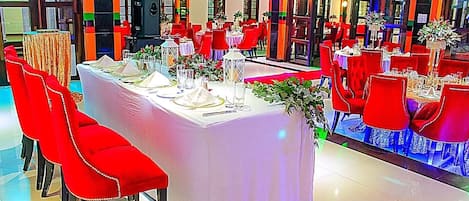 A banquet hall with red chairs and white tablecloths, featuring a stage and large windows.