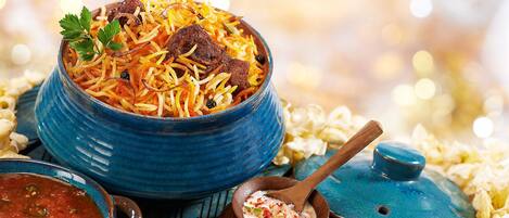 A bowl of biryani with meat and vegetables, a small bowl of sauce, and a wooden spoon.