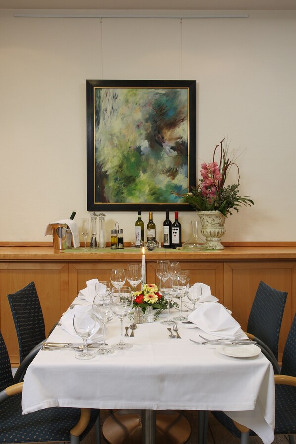A dining table set for a meal with a white tablecloth, wine bottles, glasses, and a vase with flowers. - Stadthotel Oranienburg (Oranienburg)