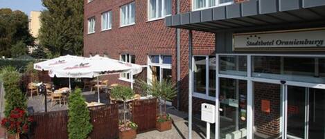 Un hôtel à la façade en briques, doté d’un espace extérieur aménagé avec des parasols et d’une enseigne indiquant « Stadthotel Oranienburg ».