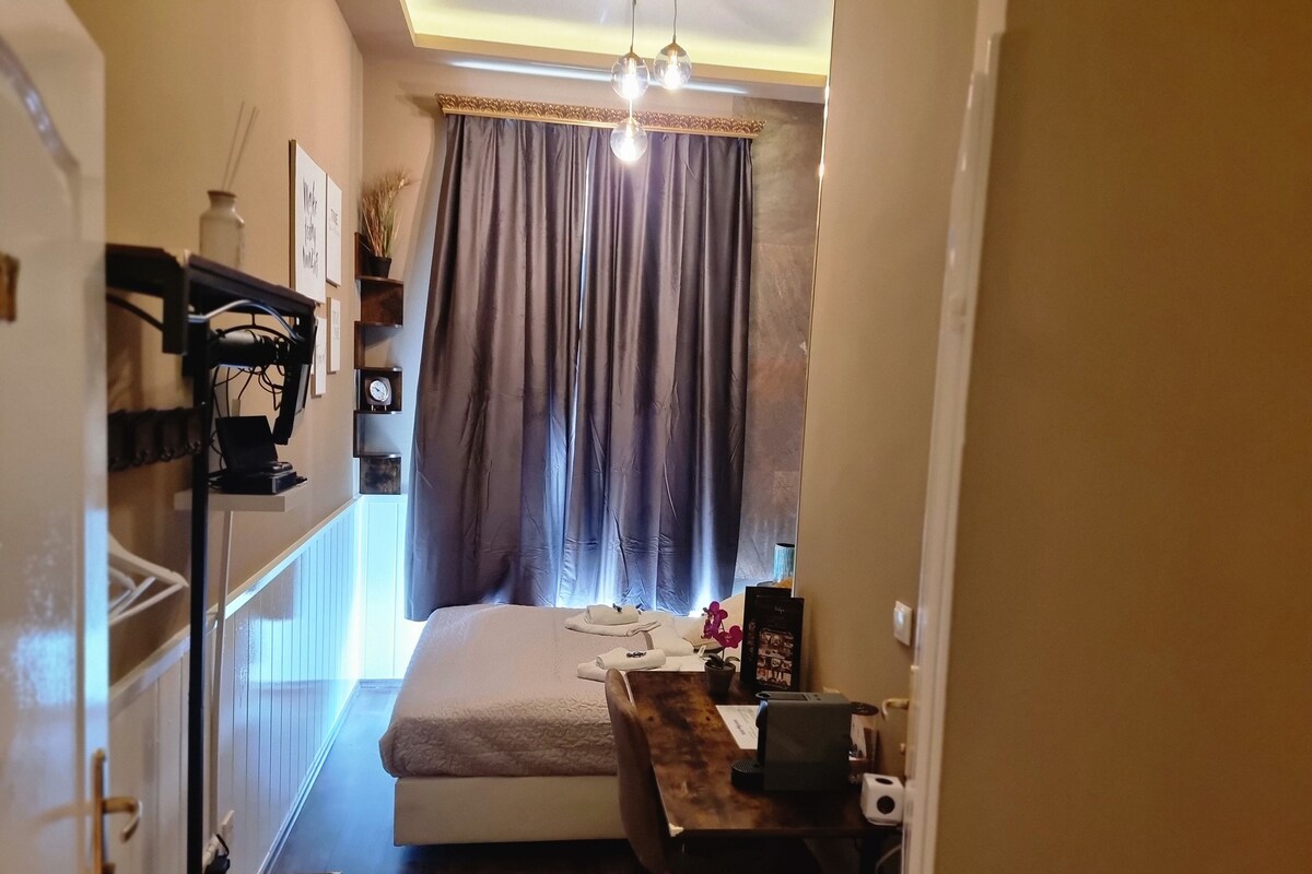 Habitación de hotel compacta con cama, escritorio y silla. Cuenta con ventana con cortinas, repisa de pared con reloj y lámpara colgante.