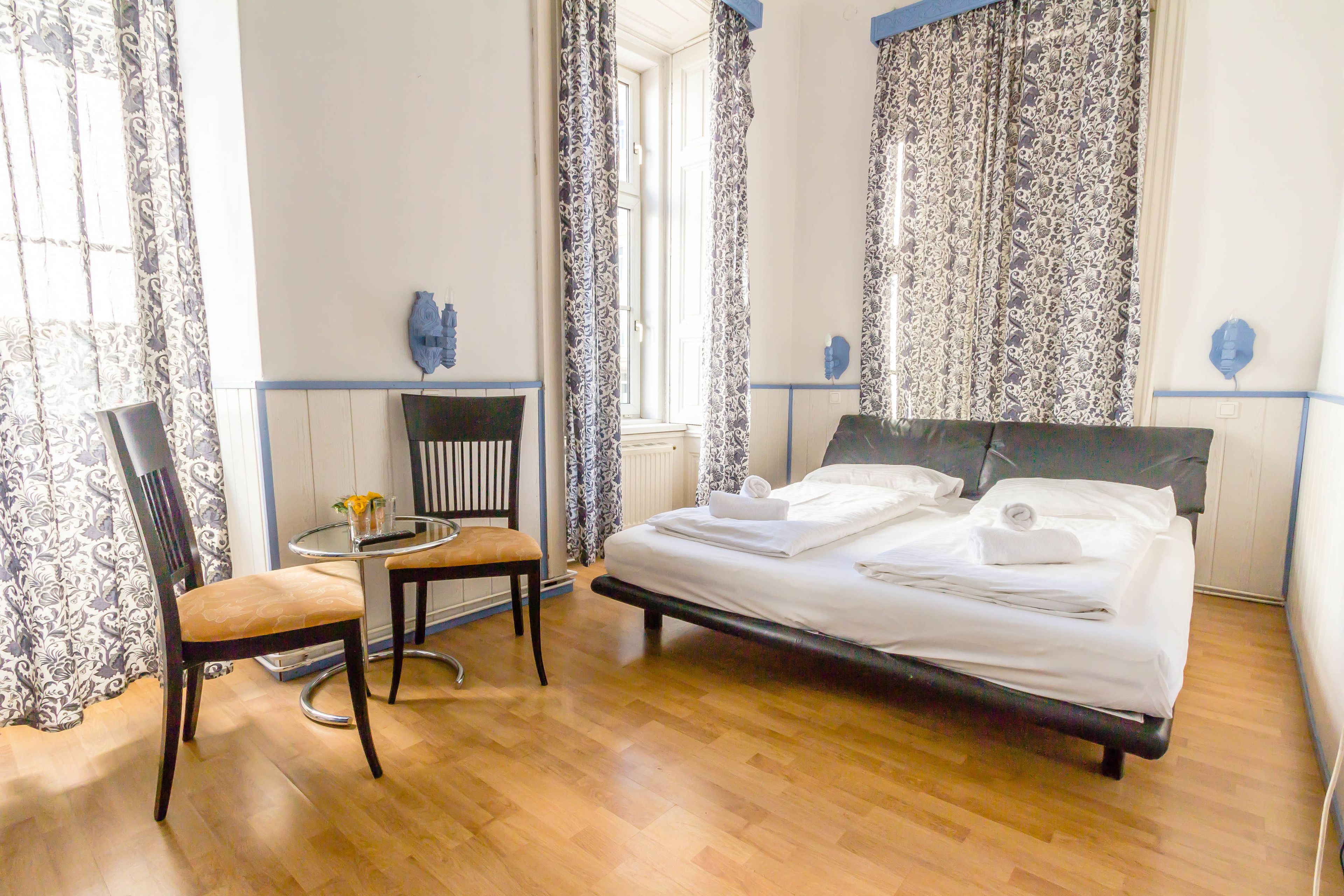 Une chambre à coucher avec un lit, une chaise, une petite table sur laquelle se trouve un vase, et des rideaux à motifs.