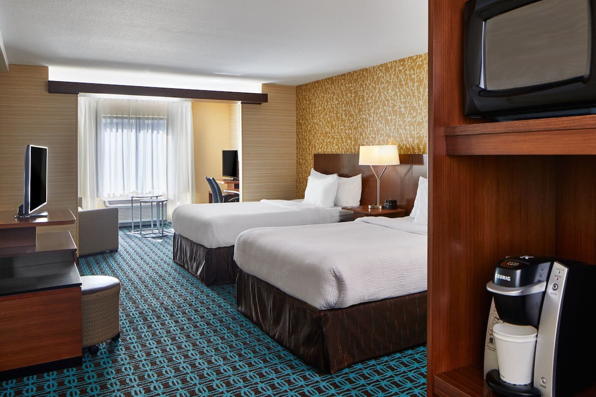 A hotel room with two beds, a desk with a flat-screen TV, a coffee maker, and a window with sheer curtains.
