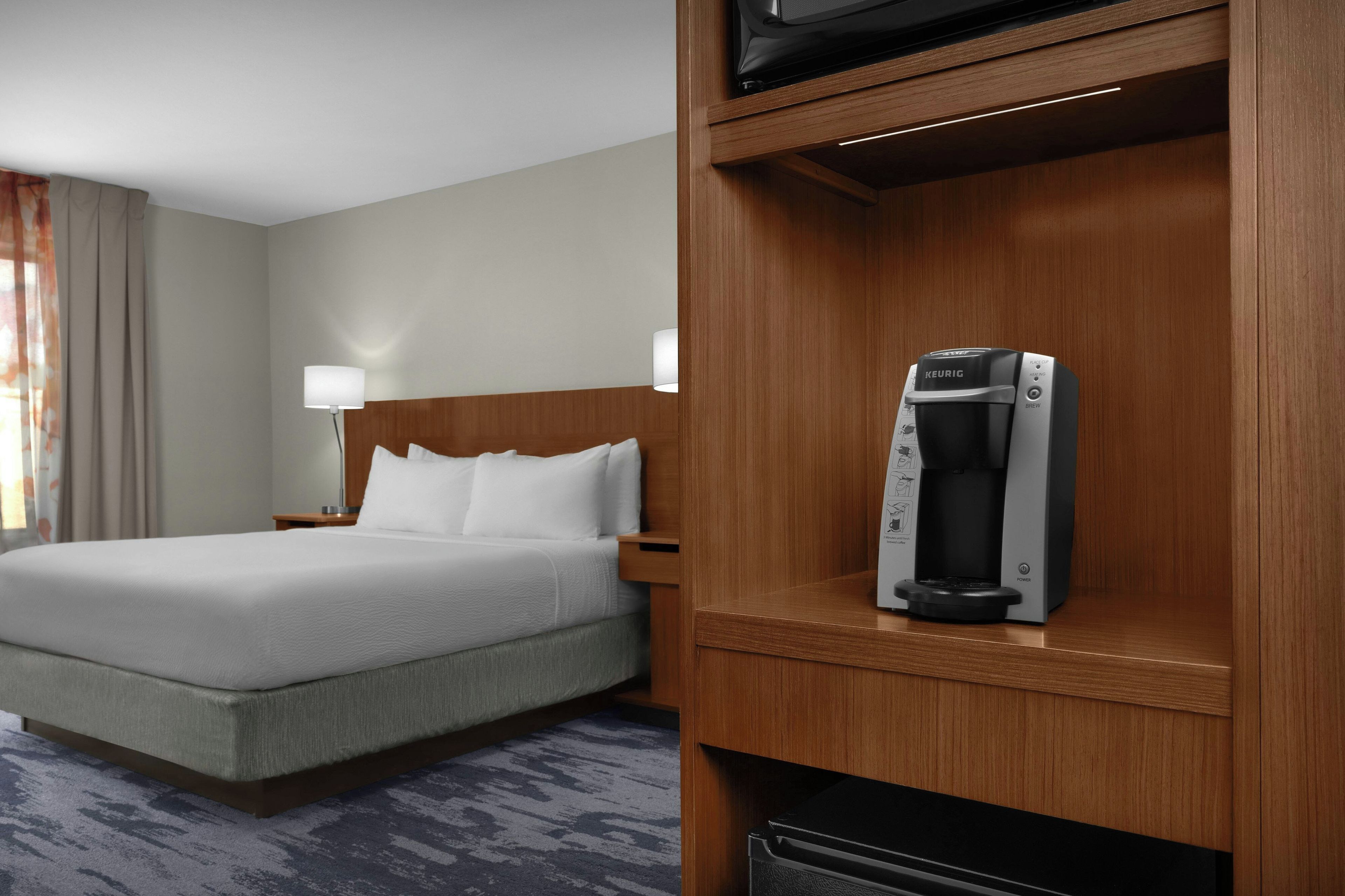 A hotel room with a bed, a nightstand, a lamp, and a Keurig coffee maker on a wooden shelf.
