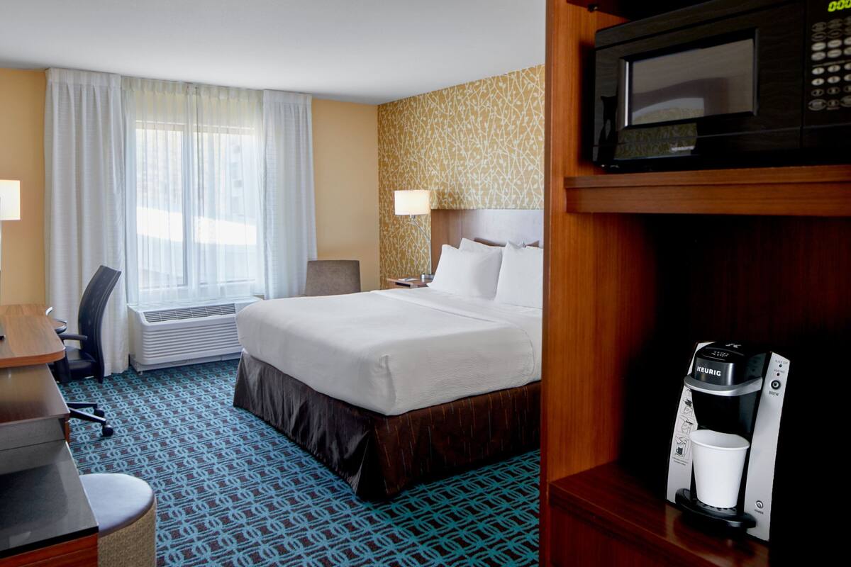 A hotel room with a bed, a desk, a chair, a coffee maker, and a window with curtains.