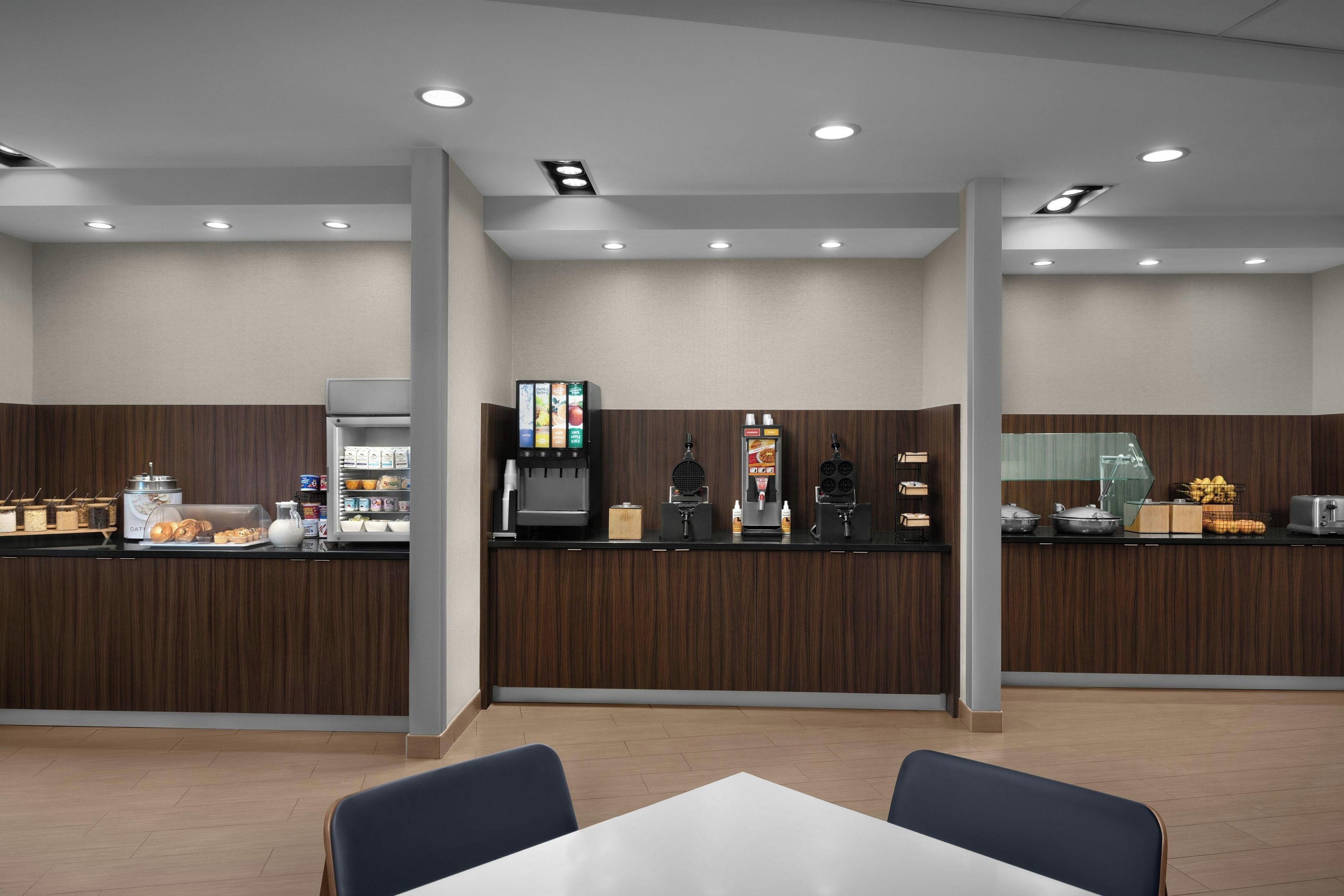 A modern cafe with a wooden counter, a coffee machine, a juice dispenser, and a variety of food items.