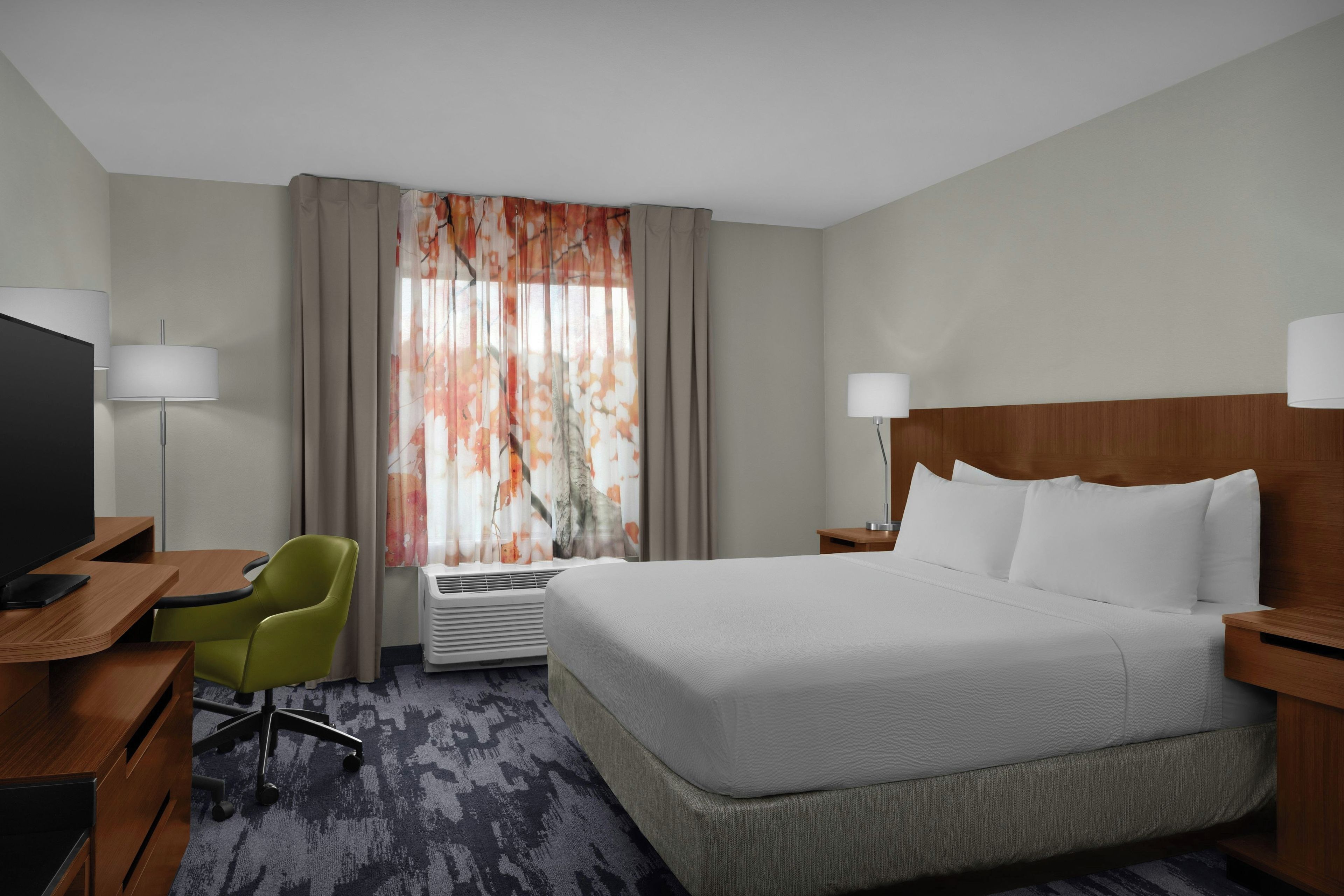 Quarto de hotel com cama, escrivaninha, cadeira, televisão e janela com cortinas.