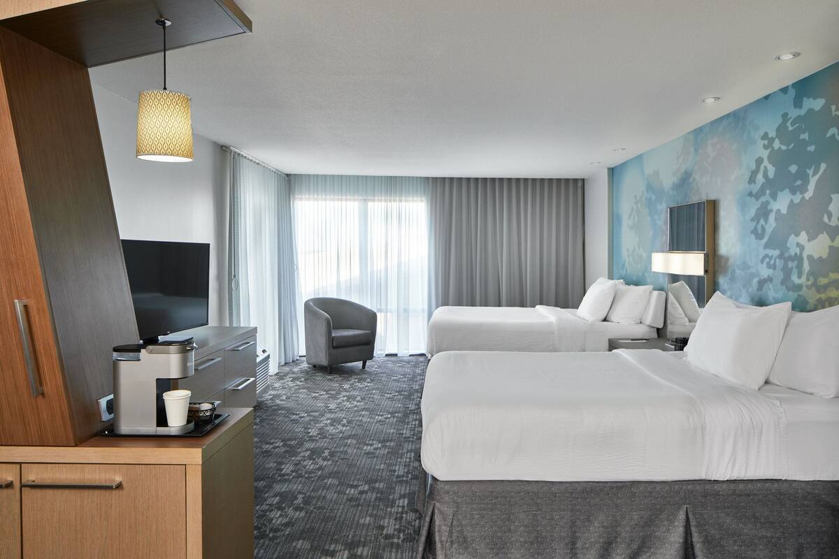A hotel room with two beds, a desk with a coffee maker, a TV, and a large window with curtains.
