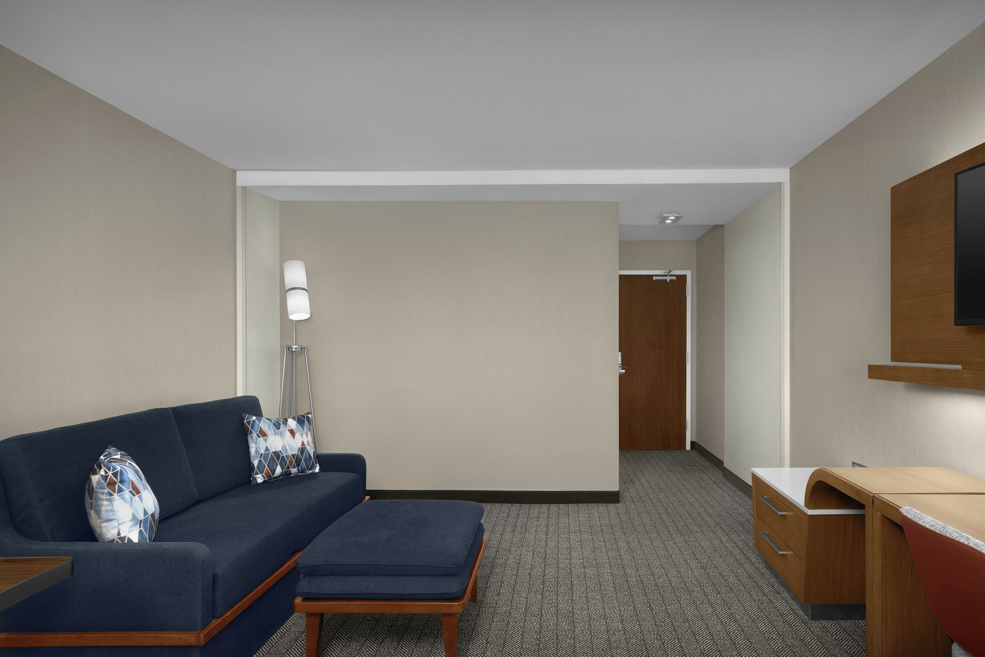 Quarto de hotel com sofá azul escuro, um apoio para os pés coordenado, escrivaninha com gavetas, luminária de pé e televisão na parede.