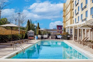 En swimmingpool med liggestole og parasoller, omgivet af en murstensmur og en bygning med mange vinduer.