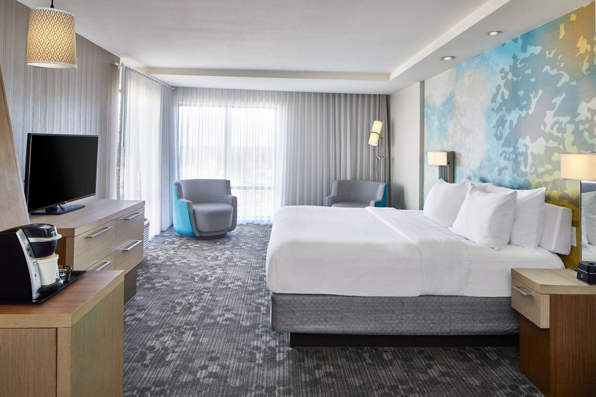 A hotel room with a large bed, a flat-screen TV, a desk with a coffee maker, and a view of the outdoors through the window.