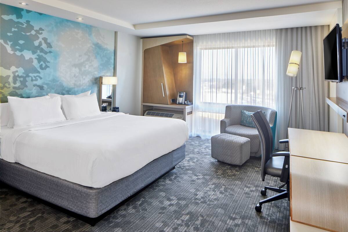 A hotel room with a large bed, a desk, a chair, a TV, and a window with curtains.