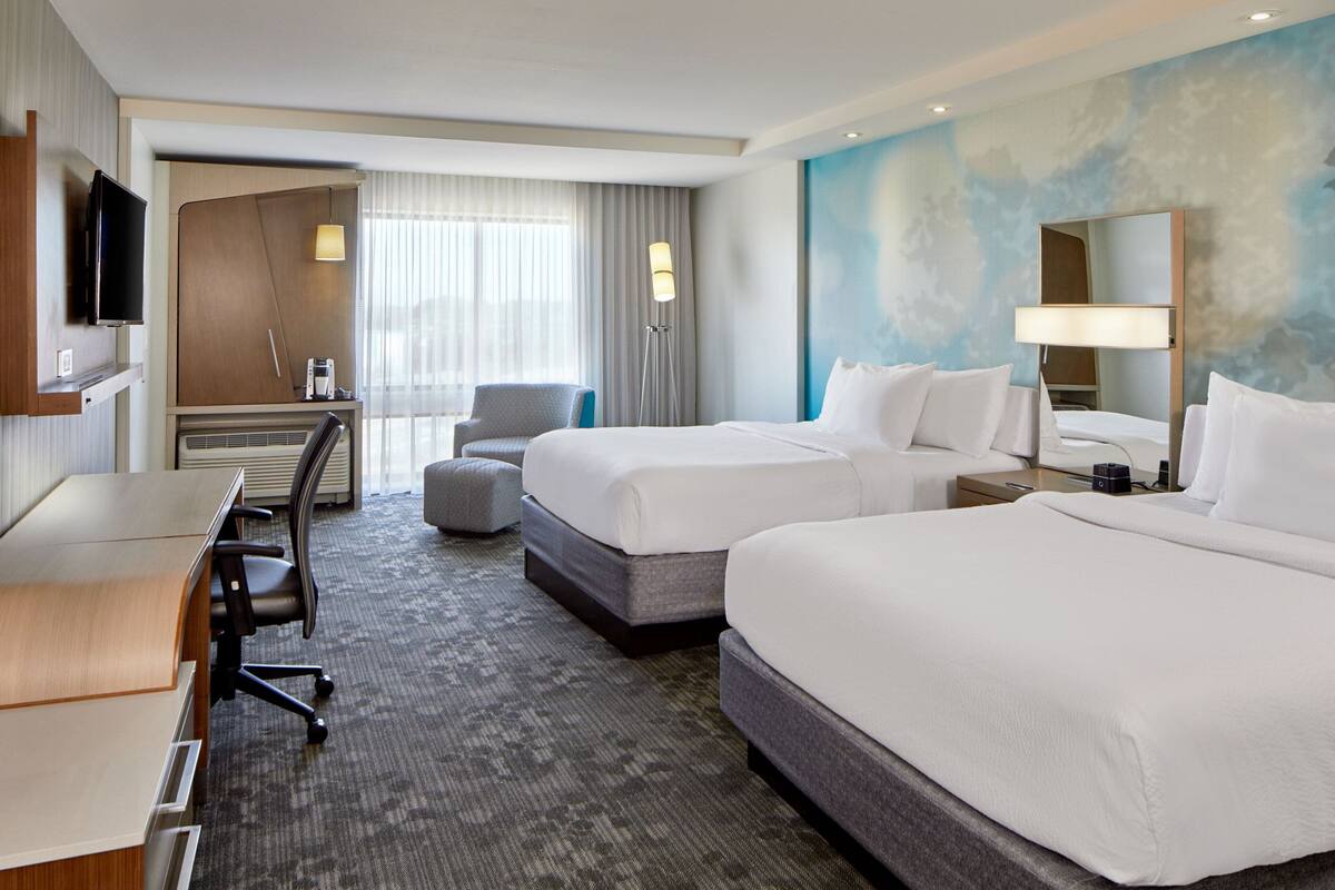 A hotel room with two beds, a desk, a chair, a TV, and a large window with curtains.